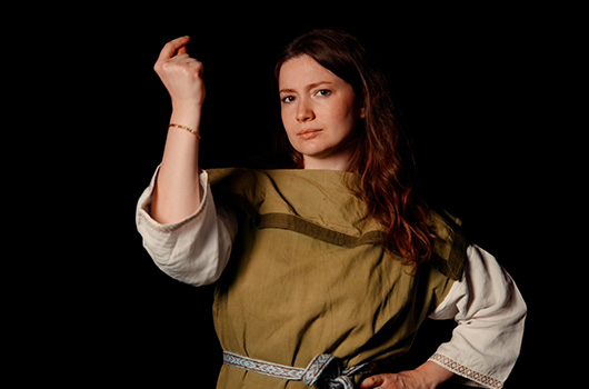 Reconstitutrice historique en tenue gauloise (tunique verte, ceinture bleue claire, sous-tunique blanche) et claquant des doigts